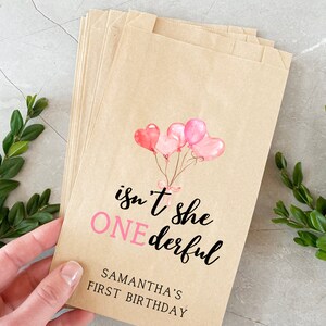 K&ouml;nnte beinhalten: Braune Papier-Geschenkbeutel mit rosa Aquarell-Ballons und dem Text "isn't she ONEderful" und "SAMANTHA'S FIRST BIRTHDAY".
