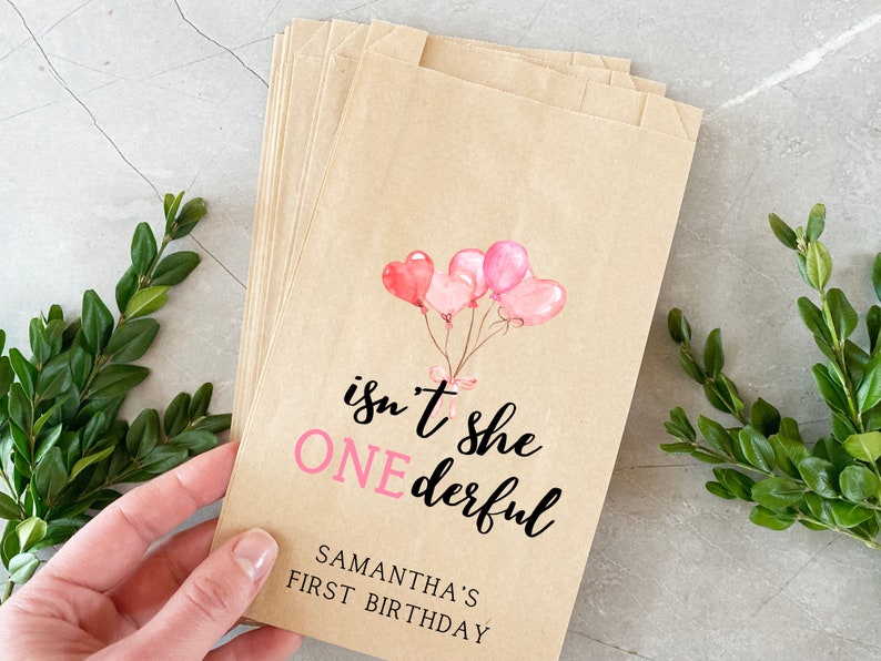 K&ouml;nnte beinhalten: Braune Papier-Geschenkbeutel mit rosa Aquarell-Ballons und dem Text "isn't she ONEderful" und "SAMANTHA'S FIRST BIRTHDAY".