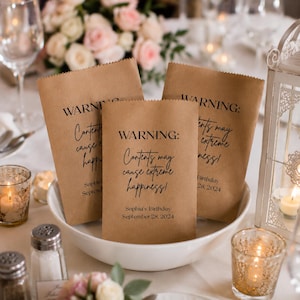 May include: Three brown paper bags with the text "WARNING: Contents may cause extreme happiness!" in black script. The bags are in a white bowl, with the text "Sophia's Birthday September 28, 2024" on one. The scene includes candles and flowers.