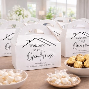 Può includere: Scatole bianche a capanna con manici, stampate con "Welcome to our Open House" in corsivo nero. Le scatole sono disposte su un tavolo di legno con ciotole di caramelle e cioccolatini incartati.
