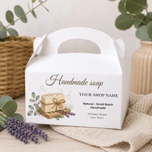 May include: A white cardboard gable box with a handle, featuring a printed label that reads Handmade soap, YOUR SHOP NAME, Natural Small Batch Handmade, and 123 Main Street Your Town, alongside an illustration of a soap bar on a wooden tray.