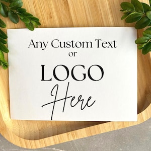 May include: A white card with the text "Any Custom Text or LOGO Here" in black, placed on a wooden tray. Green leaves are arranged around the card, adding a natural touch. The background is a light grey surface.