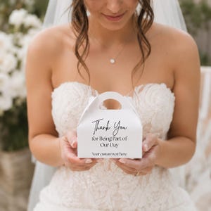 Peut inclure: Une boîte à cadeaux blanche avec une poignée, tenue par une personne portant une robe de mariée. La boîte porte l'inscription "Thank You for Being Part of Our Day", avec un espace pour un texte personnalisé en dessous.