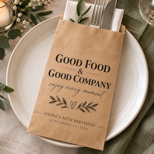 May include: A brown paper cutlery pouch containing a silver fork and knife, printed with the text "GOOD FOOD & GOOD COMPANY enjoy every moment" and "EMMA'S 40TH BIRTHDAY SEPTEMBER 12, 2026" above a graphic of two leafy branches and a small heart.