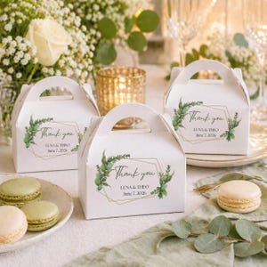 May include: White favor boxes with handles, featuring a gold geometric design and green foliage. The boxes are inscribed with "Thank you", the names "LUNA & THEO" and the date "June 7, 2026". Macarons and flowers are in the background.