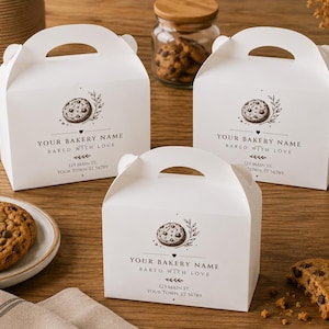Può includere: Tre scatole da pasticceria in cartone bianco con manici integrati, ognuna con una grafica stampata in nero di un biscotto con gocce di cioccolato, un rametto e il testo "YOUR BAKERY NAME BAKED WITH LOVE 123 MAIN ST. YOUR TOWN, ST 56789".