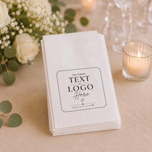 May include: A stack of white paper bags featuring a black printed label with the text Any Custom TEXT or LOGO Here, a small heart icon, and the phrase MADE WITH LOVE, framed by a decorative border on the front of the top bag.