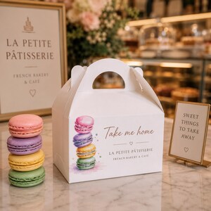 Könnte beinhalten: Eine weiße Bäckerei-Schachtel aus Karton mit Tragegriff, verziert mit einer Aquarell-Illustration von vier bunten Macarons sowie dem Text "Take me home" und "LA PETITE PÂTISSERIE FRENCH BAKERY & CAFE". Ein Stapel aus vier Macarons steht neben der Schachtel.