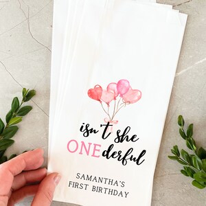 K&ouml;nnte beinhalten: Rosa und wei&szlig;e Geschenkt&uuml;ten mit einer Aquarell-Illustration von rosa Luftballons und dem Text "isn't she ONEderful" mit dem Namen Samantha's First Birthday.