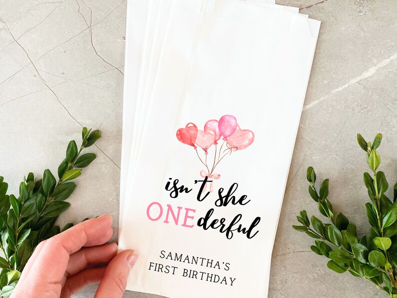 K&ouml;nnte beinhalten: Rosa und wei&szlig;e Geschenkt&uuml;ten mit einer Aquarell-Illustration von rosa Luftballons und dem Text "isn't she ONEderful" mit dem Namen Samantha's First Birthday.