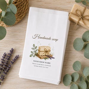 May include: A stack of white paper bags featuring a printed illustration of a bar of soap on a wooden tray with green leaves and lavender. The text reads Handmade soap, YOUR SHOP NAME, Natural, Small Batch, Handmade, and 123 Main Street, Your Town.