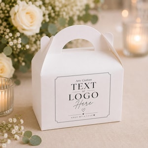 Puede incluir: Una caja de cartón blanca con asa, que presenta una etiqueta rectangular en la parte frontal. La etiqueta muestra el texto en negro: Any Custom TEXT or LOGO Here, seguido de un pequeño icono de corazón y la frase MADE WITH LOVE con corazones.