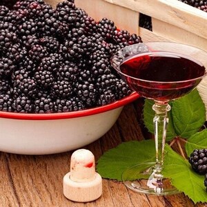 May include: A still life image featuring a bowl and a wooden crate overflowing with fresh blackberries. A glass of red liquid, a cork, and a few blackberries rest on a wooden surface, suggesting a beverage or dessert theme.