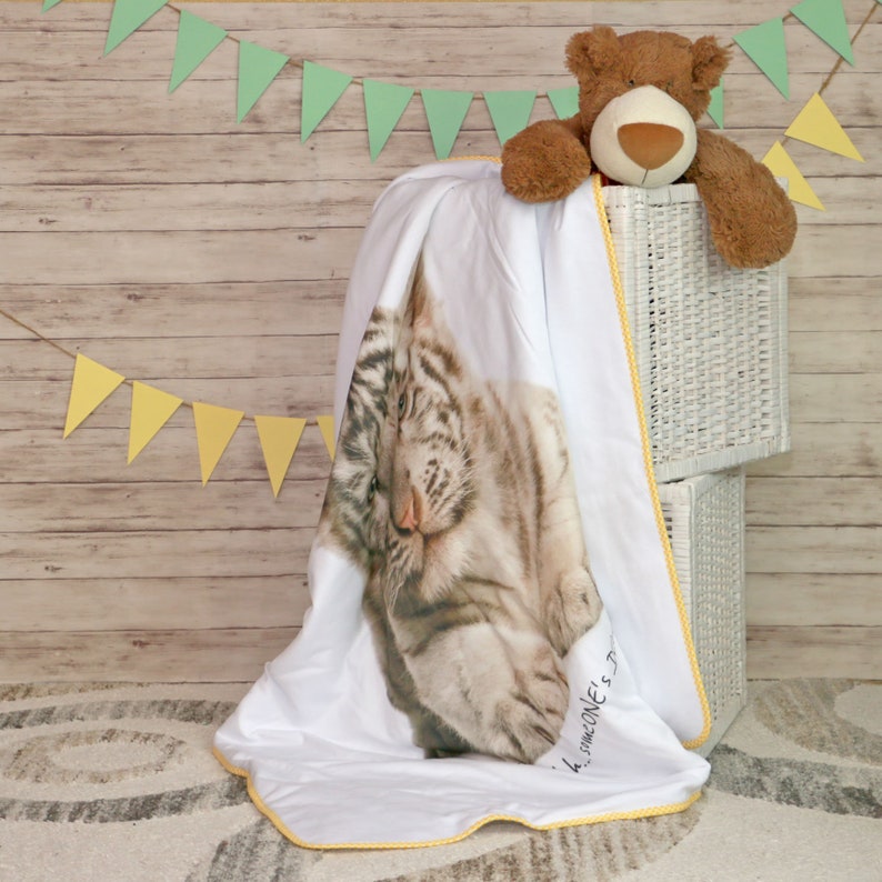 Op de afbeelding: Witte en gele babydeken met een witte tijgerprint. De deken heeft een gele rand en de woorden "Someone's Little" staan ​​gedrukt in de rechterbenedenhoek.