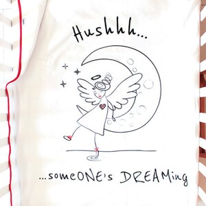 Op de afbeelding: Een witte babydeken met een rode rand. De deken heeft een zwart-witte illustratie van een engel met een halo, vleugels en een hart, die op een halve maan staat. De tekst "Hushhh... someone's DREAMING" staat onder de illustratie.