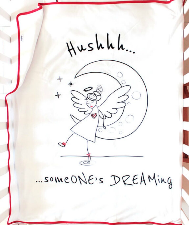 Op de afbeelding: Een witte babydeken met een rode rand. De deken heeft een zwart-witte illustratie van een engel met een halo, vleugels en een hart, die op een halve maan staat. De tekst "Hushhh... someone's DREAMING" staat onder de illustratie.