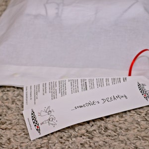 Op de afbeelding: Een witte trekkoordtas met een rood lint en een wit label met de tekst "...someone's DREAMing" en een kleine illustratie van twee engelen. Het label is met een rood lint aan de tas bevestigd.