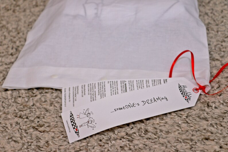 Op de afbeelding: Een witte trekkoordtas met een rood lint en een wit label met de tekst "...someone's DREAMing" en een kleine illustratie van twee engelen. Het label is met een rood lint aan de tas bevestigd.