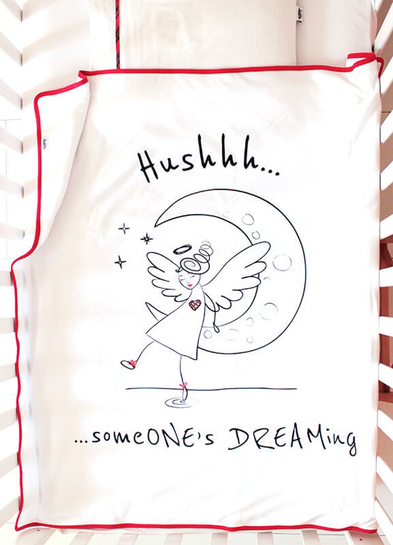 Op de afbeelding: Witte wieg lakens met een rode rand. Het laken heeft een zwart-witte illustratie van een engel met vleugels die op een halve maan staat. De tekst "Hushhh... someone's DREAMING" staat onder de illustratie.
