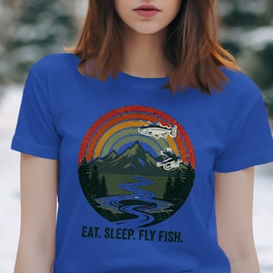 Puede incluir: Camiseta azul real con un diseño gráfico circular que muestra un paisaje montañoso, un arcoíris y peces. El texto "EAT. SLEEP. FLY FISH." está impreso debajo. La camiseta está hecha de un material suave.