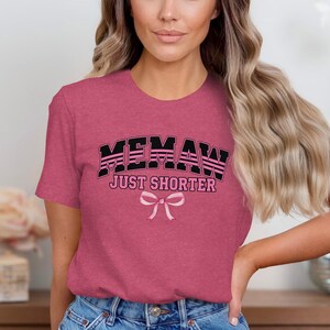 May include: A heathered mauve short-sleeve t-shirt featuring the black text MEMAW JUST SHORTER in a bold varsity font. Below the text is a light pink graphic illustration of a tied ribbon bow centered on the chest of the garment.