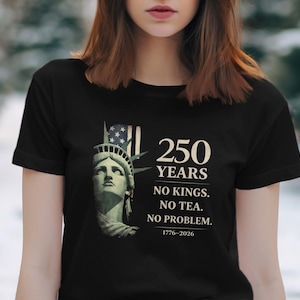 May include: Black t-shirt with a graphic of the Statue of Liberty and the text "250 YEARS. NO KINGS. NO TEA. NO PROBLEM. 1776-2026." The design includes an American flag. The shirt is a crew neck.