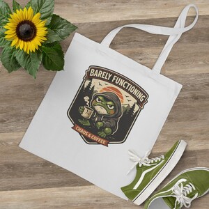 May include: White tote bag featuring a cartoon frog in a hoodie, holding a coffee cup, with the text "Barely Functioning" and "Chaos & Coffee". A sunflower and green trainers are also in the image.