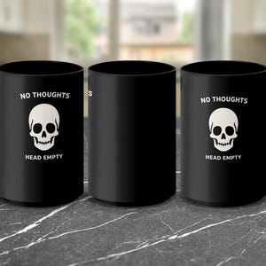 May include: Three black ceramic mugs with white text and a skull graphic. The text on the mugs reads "NO THOUGHTS" above the skull and "HEAD EMPTY" below it. The mugs are on a marble-patterned surface.