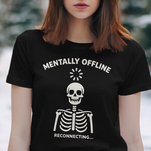 Op de afbeelding: Zwart T-shirt met de tekst "MENTALLY OFFLINE" boven een skeletafbeelding. Het skelet heeft een schedel en ribbenkast, met het woord "RECONNECTING..." eronder. Het ontwerp is wit.