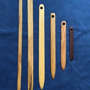 May include: Six wooden weaving shuttles of varying lengths, arranged vertically against a blue background. Each shuttle is a flat, rectangular piece of wood with a pointed end and a hole at the top. The wood tones range from light to dark.