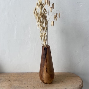 Puede incluir: Un jarrón de madera con un acabado marrón natural y una forma única e irregular. El jarrón está lleno de tallos de avena secos y cabezas de semillas.