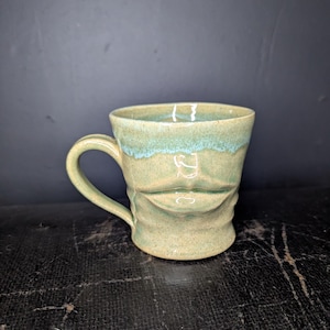 Könnte beinhalten: Eine handgefertigte Keramiktasse mit Gesichtsdesign. Die Tasse ist hauptsächlich beige mit einer türkisfarbenen Glasur am Rand. Das Gesicht hat einen geformten Mund. Der Griff ist gebogen.