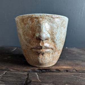 May include: A ceramic face-shaped vessel with a textured, mottled surface in shades of tan and light blue. The vessel features a detailed nose and lips, and sits on a dark wooden surface. The overall aesthetic is rustic and artistic.