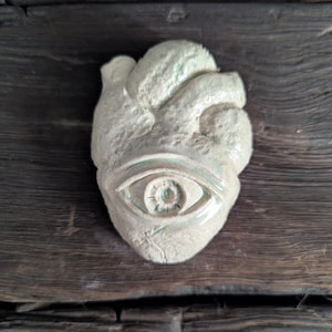 May include: A light-colored, textured sculpture of a human heart with an eye in the center. The heart is a pale, off-white color with subtle green accents. The sculpture is set against a dark, weathered wooden background.