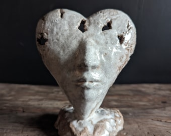 May include: White ceramic sculpture of a heart-shaped face with holes for eyes and a textured surface.