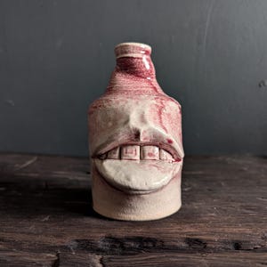 Peut inclure: Une bouteille en céramique avec un design de visage. La bouteille est principalement blanc cassé avec des accents rouges. Le visage présente une large bouche avec des dents et une langue. La bouteille est sur une surface en bois.