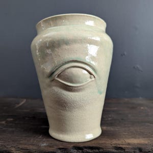 Peut inclure: Un vase en céramique blanche avec un motif d'œil vert. Le vase a une finition lisse et brillante.
