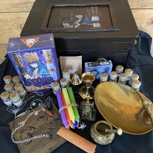 May include: A wooden box with a glass top containing a variety of items for a witch's altar. The items include candles, incense, herbs, a tarot deck, a chalice, a mortar and pestle, a small statue, and a bowl. The box is decorated with a dark wood finish and a black velvet lining.