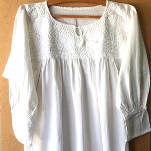 Puede incluir: Una blusa blanca de manga larga con intrincados bordados florales blancos en el pecho y los hombros. La prenda tiene un cuello redondo con una pequeña abertura central y cierre de lazo, tela fruncida bajo el canesú y mangas con puños.