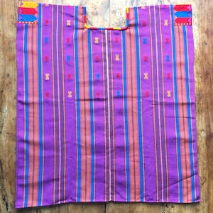 Peut inclure: Une tunique tissée violette avec des rayures verticales bleues, orange et jaunes. Le vêtement présente une encolure carrée et de petits motifs brodés rouges, bleus et jaunes dispersés sur le tissu, avec des motifs géométriques sur les épaules.