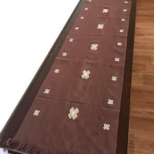 Puede incluir: Un camino de mesa largo y rectangular de color marrón con patrones geométricos bordados en blanco. El camino de mesa tiene un borde con flecos y se muestra sobre una mesa marrón oscuro. El camino de mesa está hecho de tela suave.