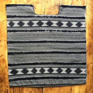 May include: A black and white woven huipil featuring a repeating diamond pattern across the top and bottom sections, interspersed with horizontal stripes of varying widths and textures throughout the rectangular garment body and neckline.