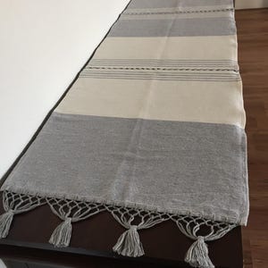 Peut inclure: Chemin de table rectangulaire à rayures crème et grises. Le chemin de table a une frange grise avec des pompons aux extrémités. Le chemin de table est posé sur une surface marron foncé.