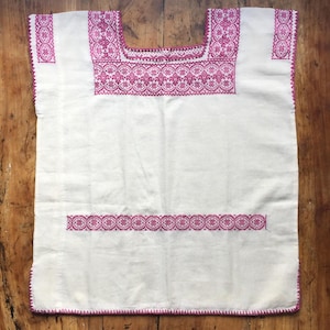 May include: A white sleeveless tunic featuring intricate magenta embroidery around the square neckline, along the shoulder panels, and in a horizontal band across the midsection, with matching magenta stitching along the bottom hemline.