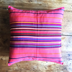 Könnte beinhalten: Ein quadratisches, pinkfarbenes Kissen mit horizontalen Streifen in verschiedenen Farben, darunter Orange, Gelb, Grün, Blau und Lila. Es liegt auf einer Holzoberfläche.