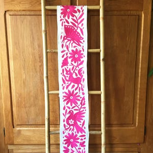Puede incluir: Un camino de mesa textil blanco con un diseño bordado en rosa vibrante. El diseño presenta una mariposa, flores, una criatura parecida a un zorro y un pájaro. El camino de mesa se muestra en una escalera de bambú contra un armario de madera.