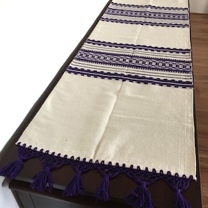 Peut inclure: Chemin de table crème avec des motifs géométriques brodés et des glands violets. Le chemin de table présente des bandes horizontales de motifs brodés et un bord crocheté avec des glands suspendus. Ce textile décoratif ajoute une touche d'élégance.