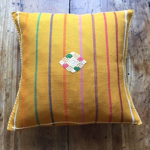 Könnte beinhalten: Senfgelbes quadratisches Kissen mit vertikalen Streifen in verschiedenen Farben. Das Kissen hat einen weiß genähten Rand und ein zentrales rautenförmiges Stickdesign mit grünen, rosa und weißen Details. Auf einer Holzoberfläche.
