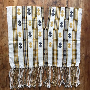 Op de afbeelding: Een witte poncho met verticale strepen in mosterdgeel en zwart, geaccentueerd met herhalende ruitvormige borduursels in dezelfde kleuren. Het kledingstuk heeft een diepe V-halsopening en een onderkant afgewerkt met lange, gedraaide franjes.