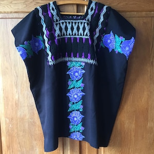 Könnte beinhalten: Eine schwarze Tunika mit aufwendigen Stickereien um den quadratischen Ausschnitt und einem vertikalen Blumenmuster in der Mitte. Das Design umfasst leuchtend violette und grüne Stickereien sowie zusätzliche Blumenmotive an Schultern und Ärmeln.